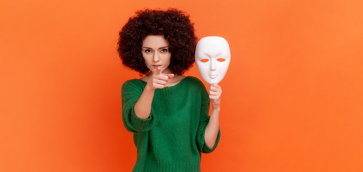 Strict bossy woman with Afro hairstyle in green casual style sweater holding white mask in hand, pointing finger to camera with serious expression. Indoor studio shot isolated on orange background.