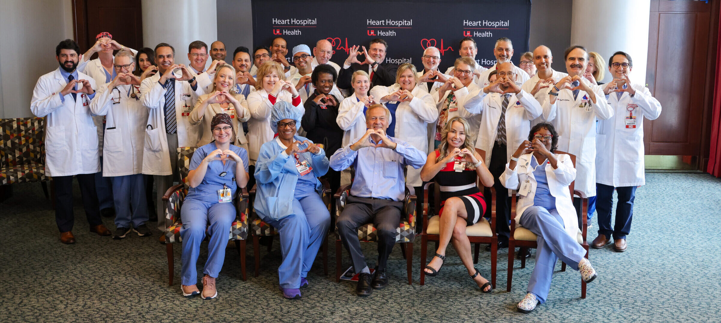 Group photo of the UofL Health – Heart Hospital team and patient John Crush. All are making a heart shape with their hands.