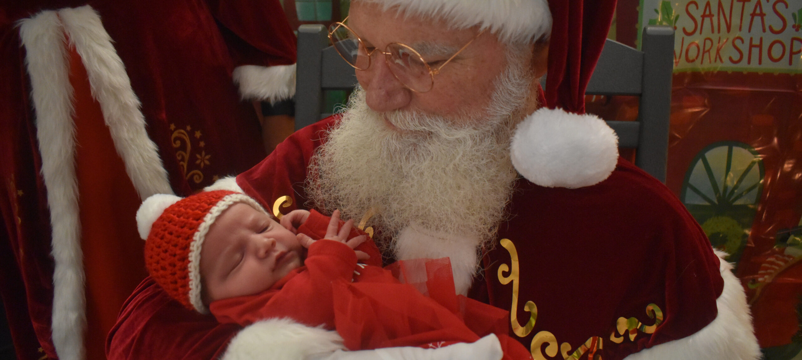 Santa holding a baby in the NICU at UofL Hospital. The baby is wearing a red Christmas-themed outfit, including a red knitted hat.