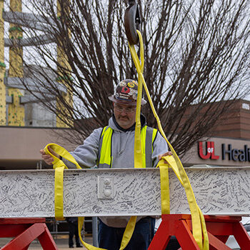 Construction worker prepares white steel beam with Bullitt County community's signatures to be hoisted in to UofL Health – South Hospital construction site.