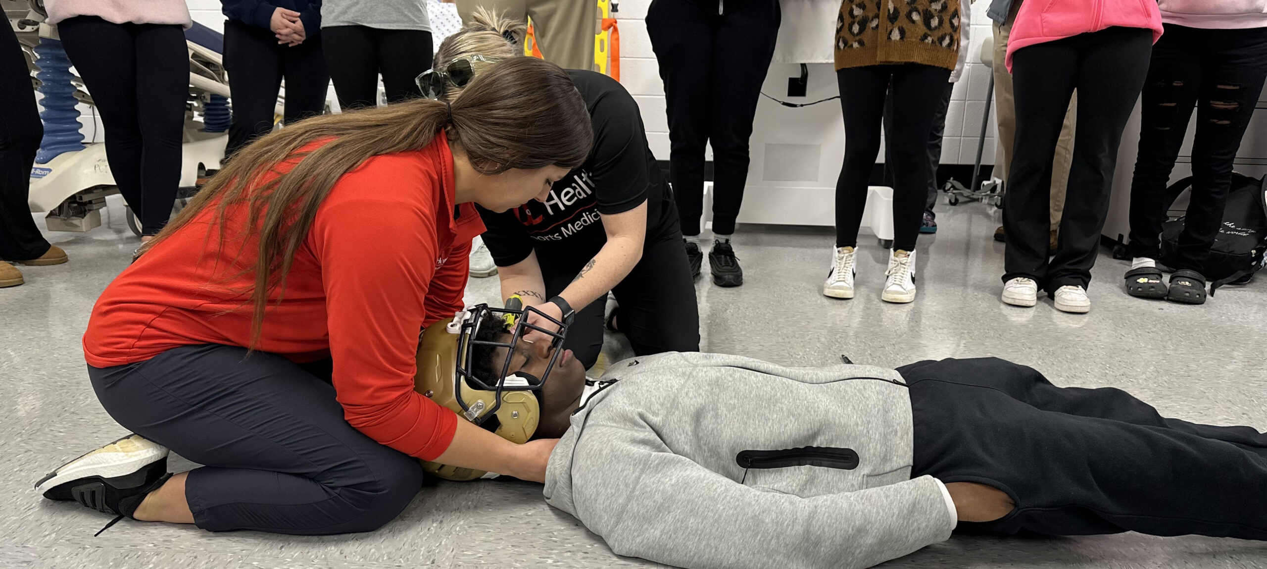UofL Health – Sports Medicine team members demonstrate how to remove a helmet from an injured football player as students at Fairdale High School observe proper athletic training techniques