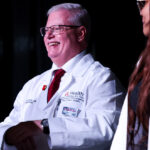 Dr. Jeffrey Bumpous standing on stage smiling in white lab coat with UofL Health and UofL School of Medicine branding. He wears a white shirt with red tie underneath.