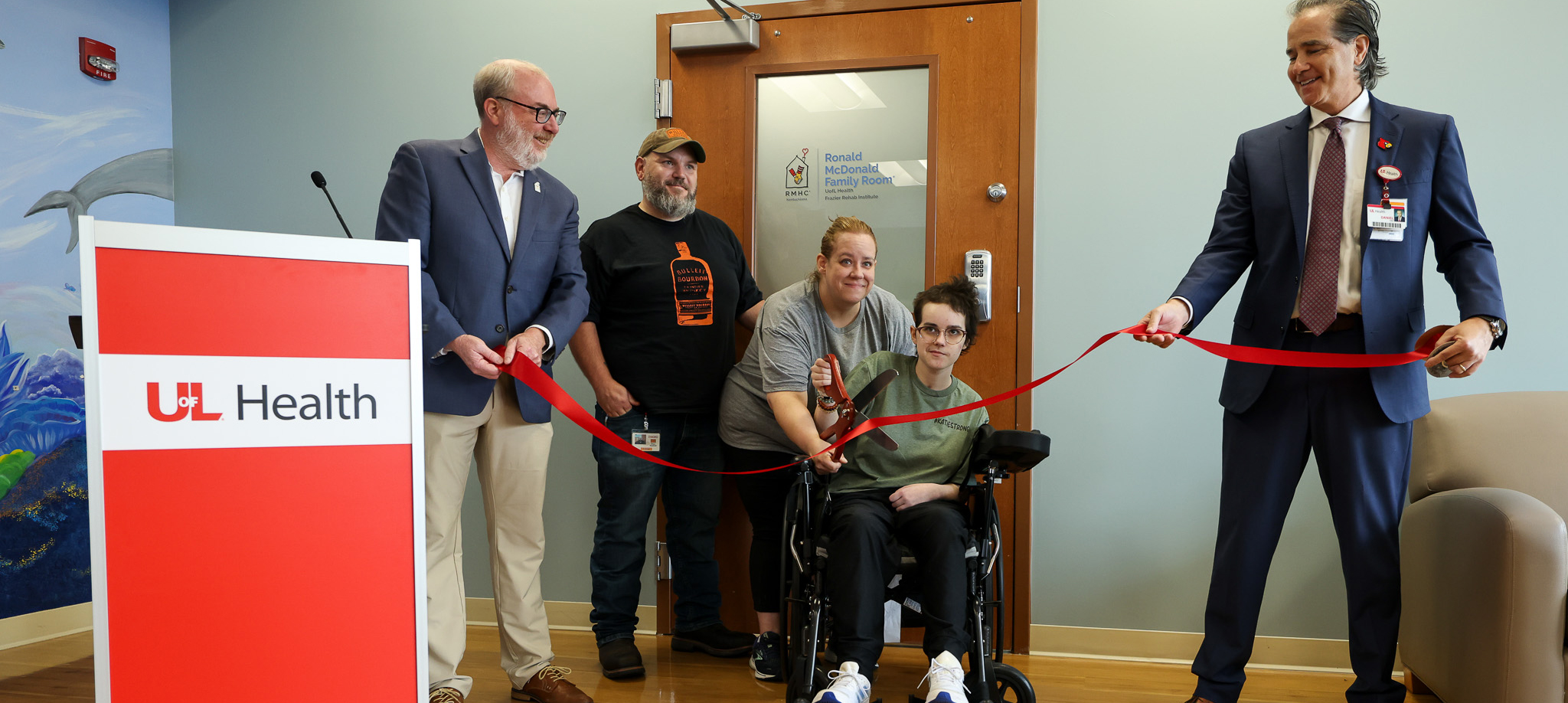 Five people participate in a ribbon-cutting ceremony for the Ronald McDonald House Charities of Kentuckiana Family Room Program at UofL Health – Frazier Rehabilitation Institute. Two individuals are standing at a podium with a UofL Health logo, while a person in a wheelchair holds scissors cutting a red ribbon, flanked by two others.