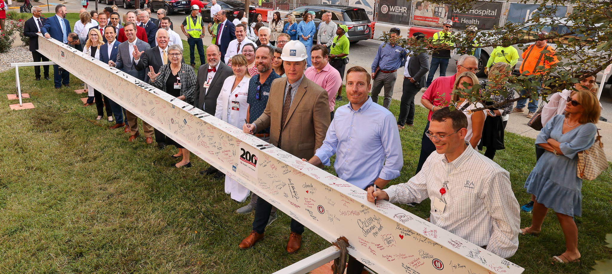 A group of people standing outdoors holding a long, horizontal beam signed by numerous individuals, with construction equipment and a hospital building in the background. The beam will be permanently placed in UofL Hospital's West Tower.