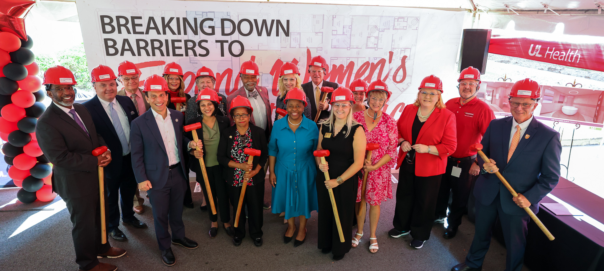 Group of individuals wearing red hard hats holding sledgehammers, standing in front of wall that reads 