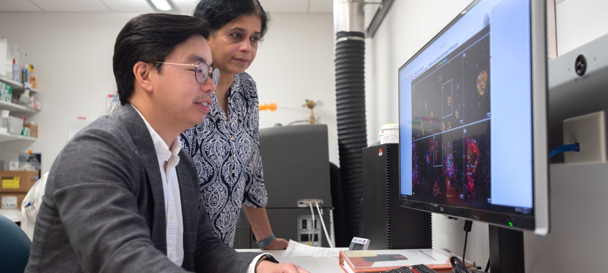 Two researchers, one seated and one standing, analyze scientific data about cancer immunotherapies on a computer screen in a laboratory setting.