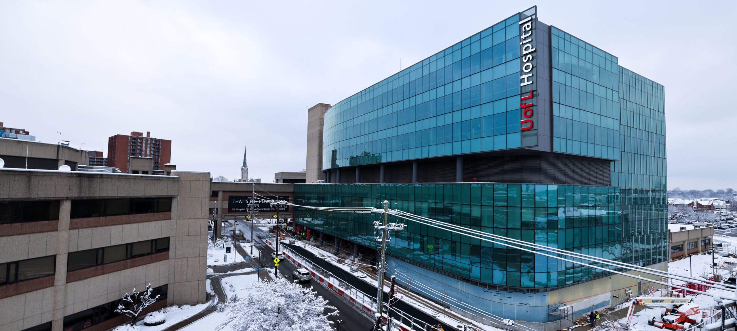 Exterior view of the completed UofL Hospital West Tower on a snowy day. The main building features a prominent glass facade, with construction and traffic visible in the foreground.
