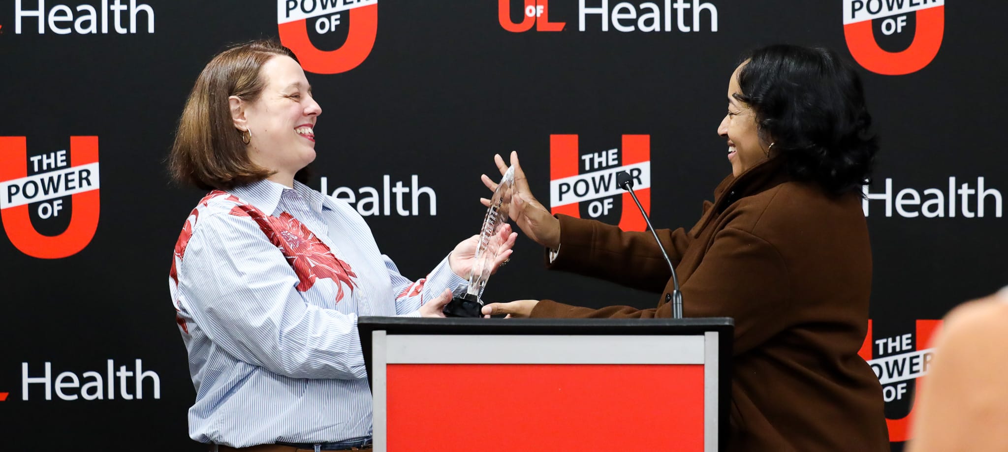Two individuals smiling and exchanging a clear Kelsie Small Future Healer award at a podium with 
