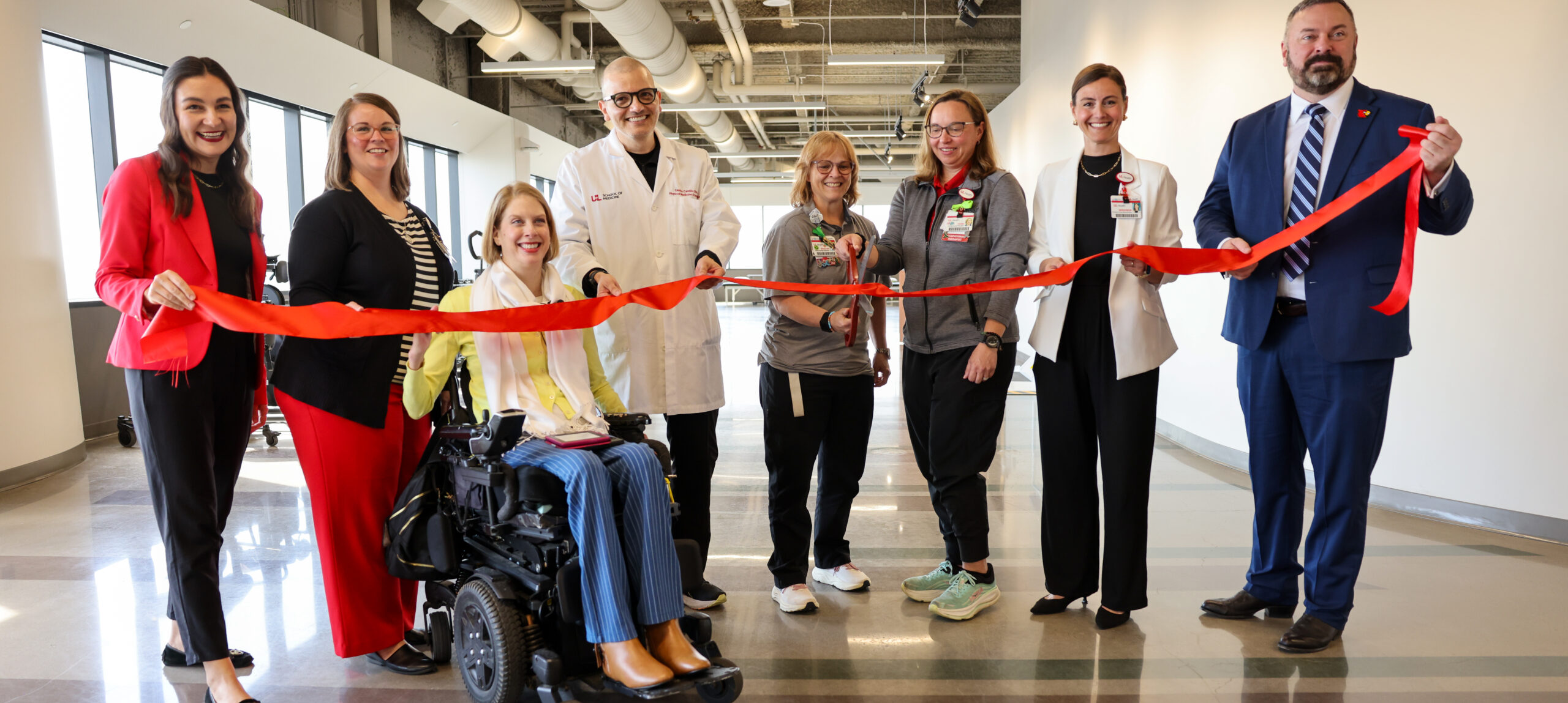 A group of eight people stand indoors, holding a red ribbon ready to be cut for an opening ceremony for the UofL Health – Frazier Rehabilitation Institute celebrated the opening of the UofL Health – Center for Advanced NeuroRehab – Assistive Technology Lab and the UofL Health – Center for Advanced NeuroRehab. One person is in a power wheelchair, holding scissors. The setting is modern, with visible ducts on the ceiling.