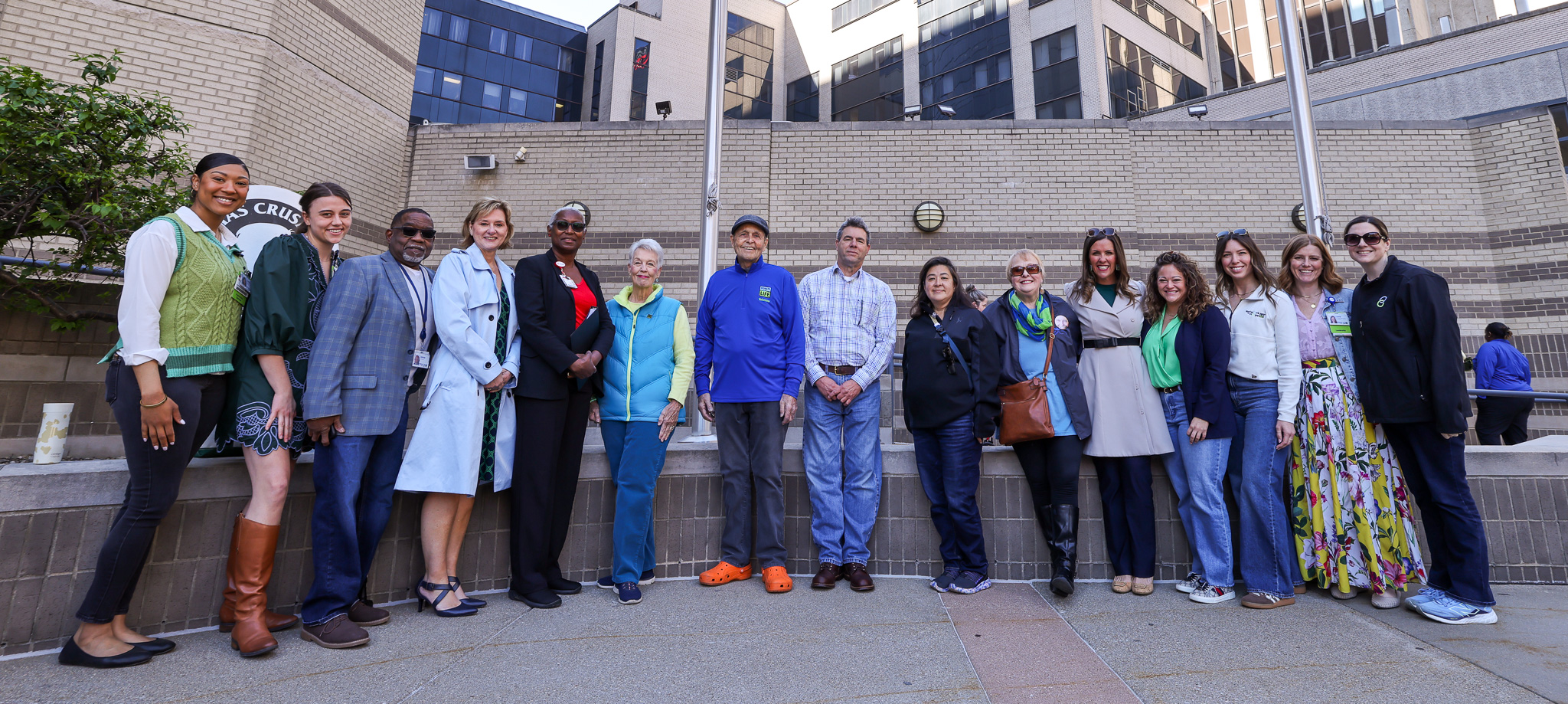 A diverse group of people stands in a line outside UofL Health – Jewish Hospital. They are positioned around a flagpole flying the American flag and a Donate Life flag. The background features a multi-story brick and glass structure with multiple windows. Some individuals hold objects, like phones or bags, and are casually dressed.